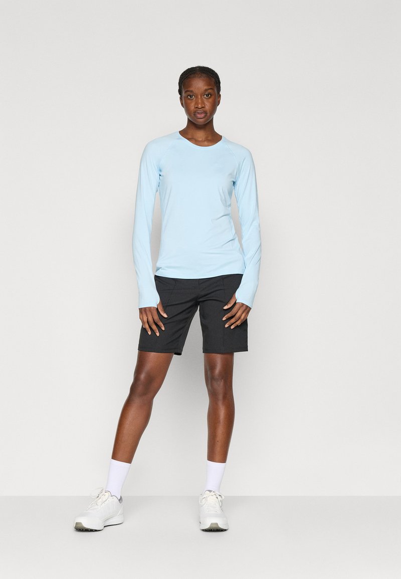 Jasnoniebieska koszula z długim rękawem w połączeniu z czarnymi szortami. Koszula ma okrągły dekolt, podczas gdy szorty sięgają kolan i mają klasyczny krój. Całość dopełniają białe sneakersy.