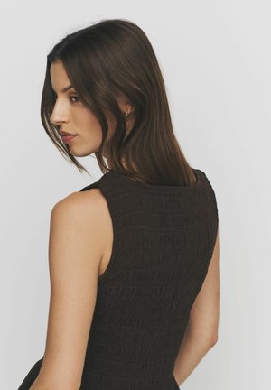 Ung kvinde med langt brunt hår iført en ærmeløs, struktureret sort top, der ser nedad mod en ensartet lys baggrund.