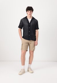 Camisa negra de manga corta con tejido de punto abierto y cuello, combinada con pantalones cortos tipo cargo beige. El modelo lleva zapatillas de deporte de color claro y calcetas blancas.