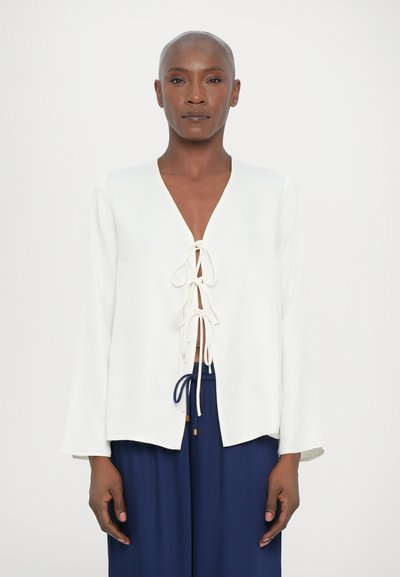 White long-sleeve top with a V-neck and front tie closure, made of smooth fabric. Worn over dark blue pants. Clean, minimal design.