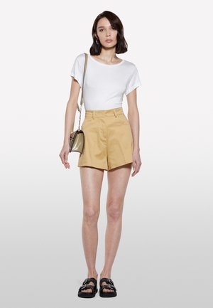 T-shirt bianco a maniche corte abbinato a un paio di pantaloni beige a vita alta. Sandali neri e una piccola borsa olive strutturata completano il look.
