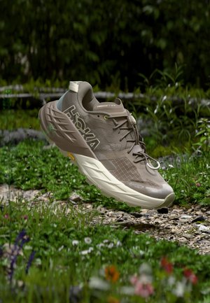 Топъл кафяв маратонка Hoka Speedgoat 7 GTX за бягане по пътеки се носи над каменна градинска пътека, заобиколена от зеленина и цветя.