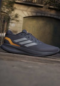 Chaussure de sport gris foncé avec trois bandes gris clair et un accent moutarde, posée sur une surface pavée avec un arrière-plan urbain flou.
