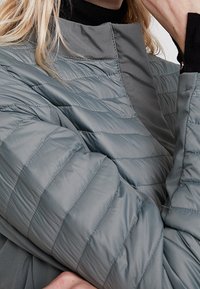 Primo piano di una persona che indossa una giacca trapuntata grigia chiaro con cuciture orizzontali sopra un maglione a dolcevita nero.