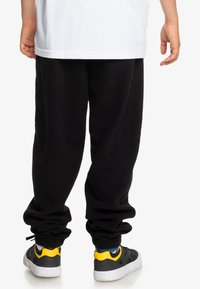 Svarta fleecebyxor med elastisk midja och mudd, som kombineras med grå sneakers med gula detaljer och vita sulor.