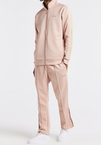 Světlé růžové teplákové souprava z hladké látky; bunda na zip s vysokým límcem, postranní pruhy a odpovídající kalhoty s větracími otvory na zip u kotníků.