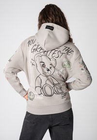 Grijze hoodie met een berenafbeelding op de achterkant, voorzien van zwarte lijntekeningen en kleurrijke tekst. Het beschikt over een capuchon met trekkoord en geribde manchetten.