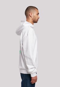 Felpa bianca con cappuccio e tasca a marsupio, polsini a coste e un dettaglio grafico verde sulla parte posteriore. Indossata con jeans in denim blu.