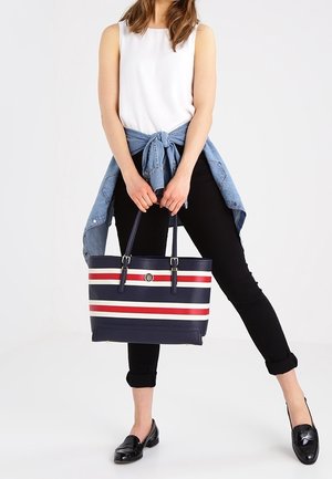 Kobieta ubrana w biały top bez rękawów, czarne spodnie, niebieską koszulę zawiązaną w pasie, trzyma granatową torbę typu tote z czerwono-białymi poziomymi paskami.
