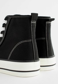 Schwarze High-Top-Sneaker aus Canvas mit weißen Schnürsenkeln, kontrastierendem Stich und Gummisohlen, die an der Basis mit einem schwarz-weißen gestreiften Akzent versehen sind.