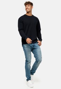Schwarzes Langarm-T-Shirt aus strukturiertem Stoff, kombiniert mit hellblauen, lässig verwaschenen Jeans und weißen Sneakers. Das Model steht in einer dynamischen Pose.