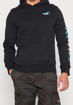 Mann trägt schwarzen Kapuzenpullover mit blauem Logo auf Brust und Text auf dem Ärmel, kombiniert mit dunkler Camouflage-Cargohose, Hände entspannt an den Seiten.