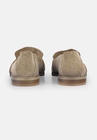 Zapatos de ante beige con un trasero redondeado, tacón de madera bajo y detalles perforados a lo largo del borde superior. Textura suave y diseño minimalista.