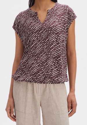 Kobieta w bluzce z krótkim rękawem w bordowo-biały wzór z dekoltem w serek oraz beżowych spodniach z wysokim stanem.