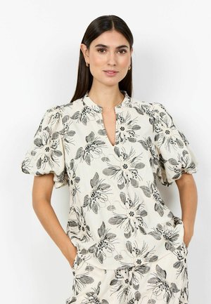 Vrouw met lang donker haar draagt een bijpassende crème outfit met zwarte bloemenprint, opgeblazen korte mouwen en handen in de zakken tegen een witte achtergrond.