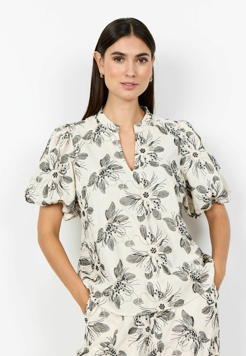 Femme aux longs cheveux foncés portant une tenue crème assortie avec un imprimé floral noir, des manches courtes bouffantes, et les mains dans les poches sur fond blanc.