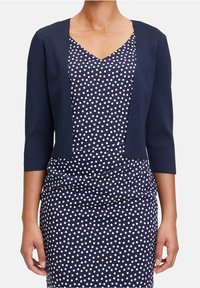 Marinblå kort cardigan med trekvartslånga ärmar, bärs över en prickig klänning. Klänningen har en v-ringning och rynkade detaljer.
