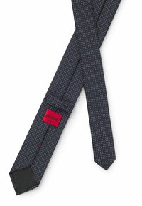 Cravatta blu con pois bianchi, forma stretta, texture del tessuto, presenta un'etichetta rossa con "HUGO" e l'etichetta "MADE IN ITALY", design elegante.