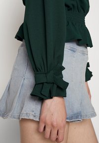 Grüne Bluse mit Rüschensäumen, die mit Schleifen gebunden sind, kombiniert mit einem hellen Jeans-Minirock mit ausgestelltem Schnitt und ausgefranstem Saum.