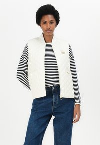 Beige gewatteerde vest met een hoge kraag en gouden details, gedragen over een zwart-wit gestreept shirt, gecombineerd met blauwe high-waisted jeans.