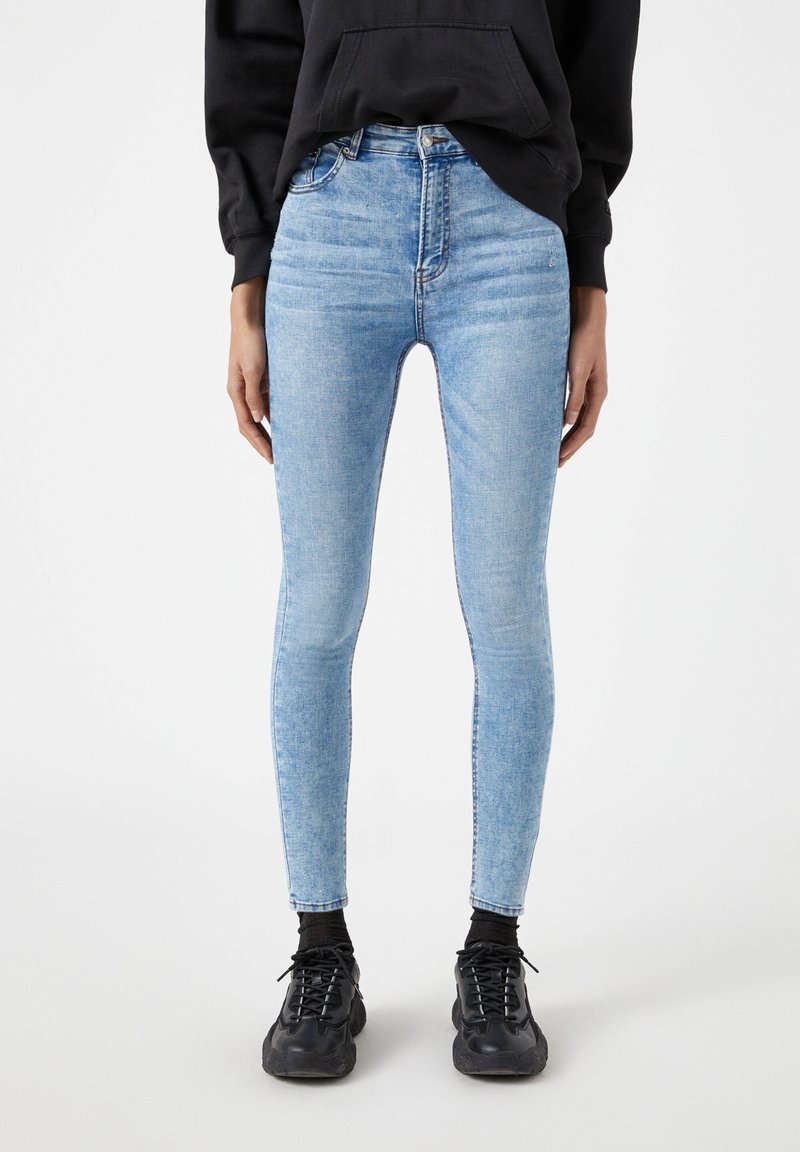 Jean skinny bleu clair à taille haute, présentant un délavage subtil, cinq poches et une fermeture éclair. Associé à des baskets noires.