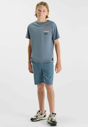 Adolescent blond debout, portant un t-shirt graphique bleu, un short bleu et des baskets noires et blanches sur un fond blanc.