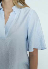 Chemise bleu clair à manches courtes avec une coupe décontractée, en tissu texturé et un col en V. Comprend un col à boutons et un drapé doux.
