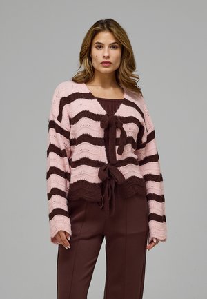 Mujer con un cárdigan de punto a rayas rosas y marrones con cintas sobre pantalones marrones, sobre un fondo gris liso.
