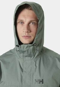 Chaqueta impermeable de color verde claro con capucha, que cuenta con toggles ajustables y un logo negro en el pecho. Textura suave y diseño sencillo.