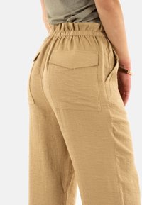 Pantalons en lin beiges avec une taille élastique et deux poches arrière. Le tissu a une finition texturée avec une coupe décontractée et une jambe large.