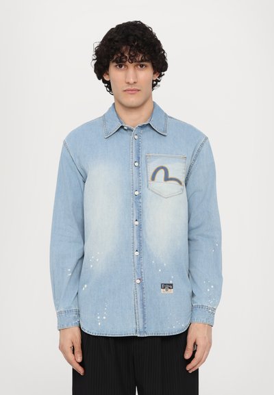 Uomo con capelli ricci e scuri indossa una camicia di jeans azzurro chiaro con bottoni, tasca frontale e dettagli di schizzi di vernice, in piedi contro uno sfondo semplice.