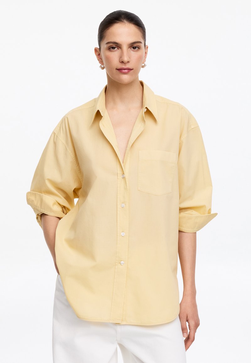 Femme aux cheveux foncés attachés, portant une chemise ample jaune pâle à boutons avec les manches retroussées et un pantalon blanc, debout devant un fond blanc.