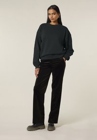 Sweatshirt noir surdimensionné avec des poignets côtelés, associé à un pantalon en velours côtelé large de couleur foncée et des chaussures à plateforme noires et épaisses. Le modèle pose avec une main dans sa poche.