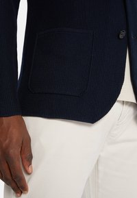 Námořnický textured blazer s klopovým límcem, na jedno knoflík a s náprsní kapsou, kombinovaný se světlými kalhotami.