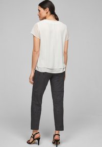 Blusa bianca leggera e semitrasparente con maniche corte, schiena a strati, abbinata a pantaloni aderenti grigi scuri e tacchi neri appuntiti.