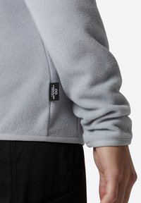 Sudadera de forro polar gris claro con una textura sutil, que presenta una etiqueta de marca en el lateral y un dobladillo oscuro de contraste.