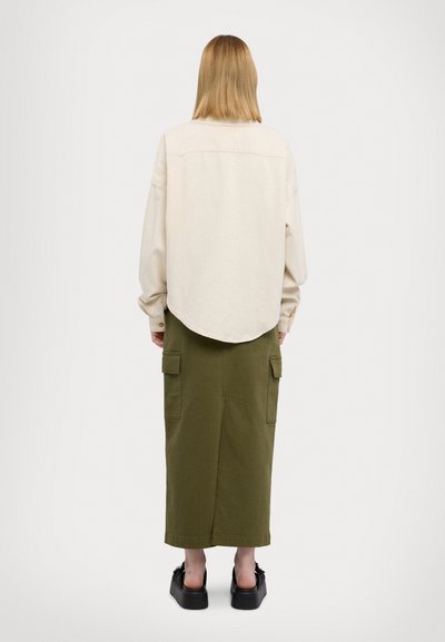 Camisa oversize color crema con dobladillo curvado, combinada con una falda larga de color verde oliva que cuenta con bolsillos tipo cargo. Sandalias de plataforma negras completan el look.