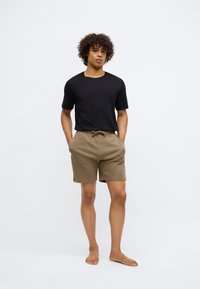 Tânăr stând desculț cu mâinile în buzunare, purtând un tricou negru și pantaloni scurți cu șnur maroniu, pe un fundal alb simplu.