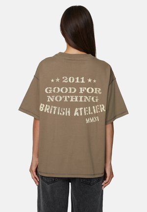 Donna con lunghi capelli scuri che indossa una maglietta marrone larga con testo bianco "2011 GOOD FOR NOTHING BRITISH ATELIER" sul retro.