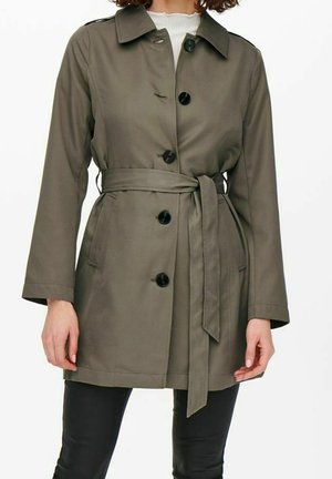 Femme portant un trench-coat ceinturé vert olive longueur genou avec des boutons noirs, associé à un pantalon noir et un haut blanc en dessous.