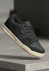 Black sneaker with perforated toe, smooth leather upper, white sole, and gum rubber outsole. Features a logo tag on the side.