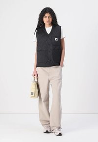 Schwarz gesteppte Weste mit runden Mustern, vier Fronttaschen und einem Logo-Patch. Kombiniert mit hellbeigen Hosen und einer kleinen hellbeigen Tasche.