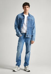Chaqueta de mezclilla y vaqueros de un azul claro, ambos con un lavado desgastado, combinados con una camiseta blanca y zapatillas gris. Detalles de costura visibles en primer plano.