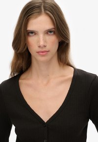 Joven mujer con cabello castaño claro y pecas, vistiendo una blusa negra de canalé con escote en V y botones, frente a un fondo liso.