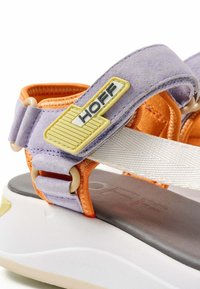 Sandal de color púrpura y naranja con suela plana. Presenta correas ajustables, acentos texturizados y un detalle de logo. Materiales de ante suave y tela.