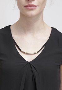 Svart V-hals topp med en plisserad design, med ett böjt metallackcent på ett svart snöre halsband. Slät tygtextur, minimalistisk detaljering.