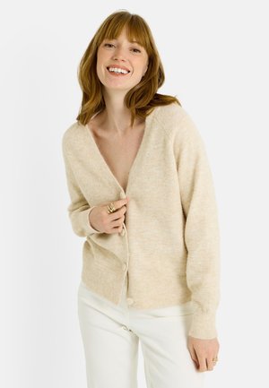 Cárdigan de punto beige con escote en V y cierre de botones, con mangas largas y textura suave, combinado con pantalones blancos.