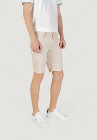 Pantaloni corti beige con un design testurizzato, tasche e zip, abbinati a sneakers bianche e una polo bianca con dettagli neri.