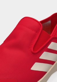 Κοντινή λήψη ενός κόκκινου αθλητικού παπουτσιού χωρίς κορδόνια με λευκές ρίγες και υφασμάτινο επάνω μέρος με υφή, που διαθέτει μαλακό πέλμα στον αστράγαλο και μπεζ σόλα.