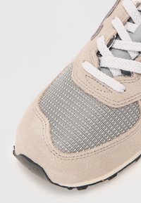 Gros plan de l'avant d'une sneaker beige et grise avec des textures en daim et en maille, des lacets blancs et une semelle à bout noir sur un fond blanc.